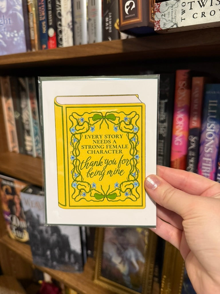 Hand holding a card with a yellow book design in front of a bookshelf.