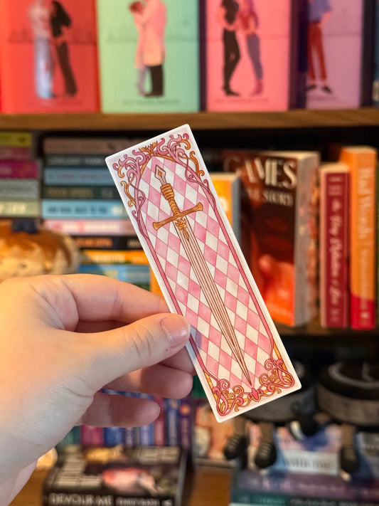 Hand holding a decorative bookmark with checkered pattern and sword design, against a bookshelf background.