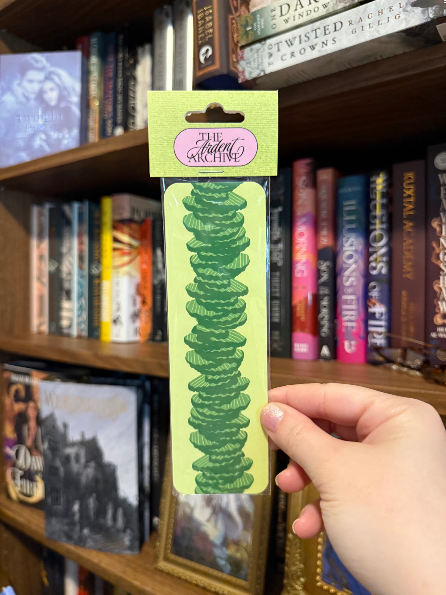 Hand holding a green pickle bookmark with 'The Ardent Archive' branding in front of a bookshelf.