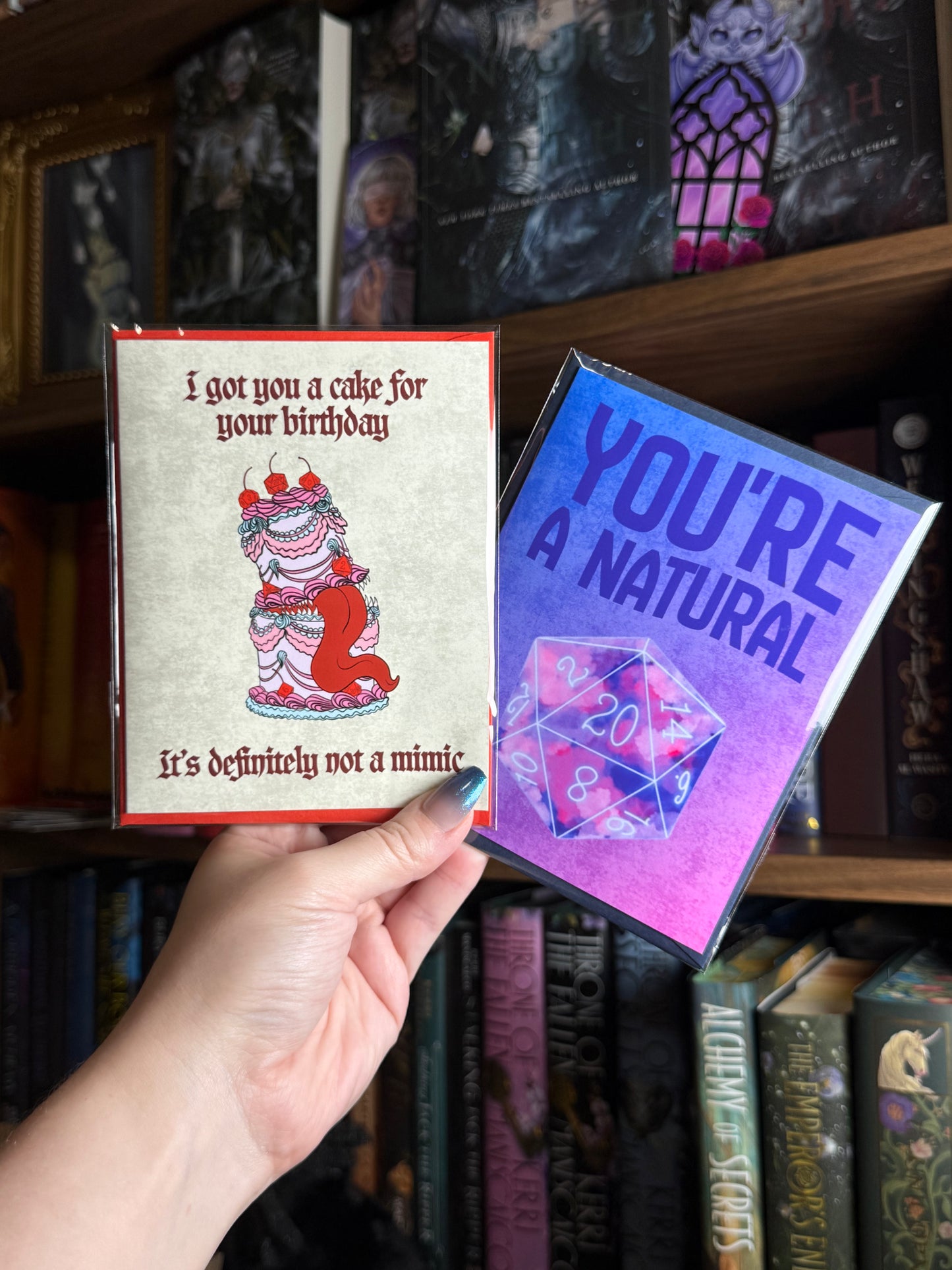 A hand holds two themed greeting cards in front of a bookshelf. The left card has a beige background with a red border and an illustration of a cake-like figure, with text that reads 'I got you a cake for your birthday / it's definitely not a mimic'. and the right side says "you're a natural" with a D20 die.