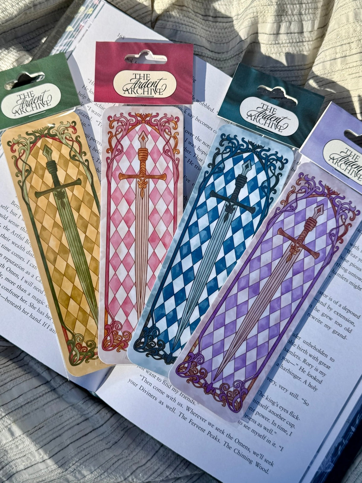 Four decorative paper bookmarks with swords in sleeves, each in a different color: yellow, pink, blue, and purple. They are arranged on top of open book pages outdoors.