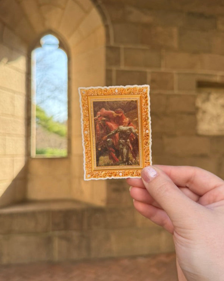 Hand holding a small framed artwork in front of a stone wall with an arched window.