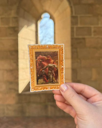 Hand holding a small framed artwork in front of a stone archway.