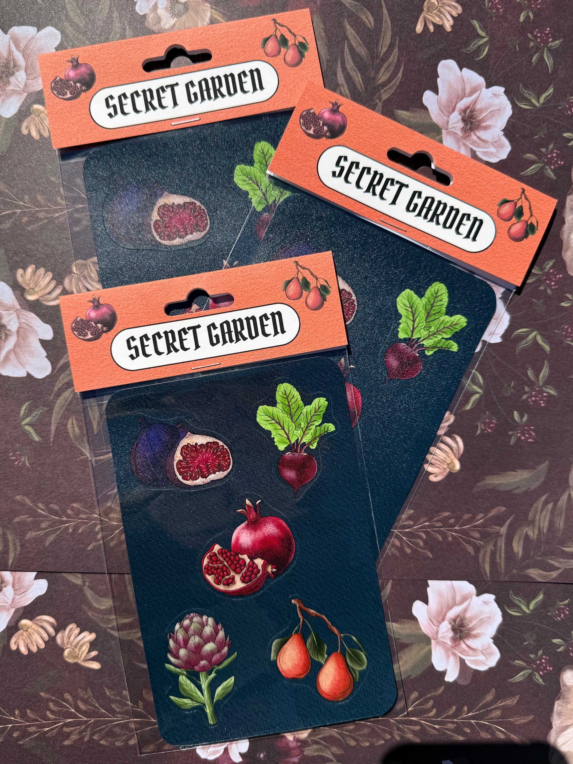 Three packages of 'Secret Garden' stickers with illustrations of fruits and vegetables on a floral background.