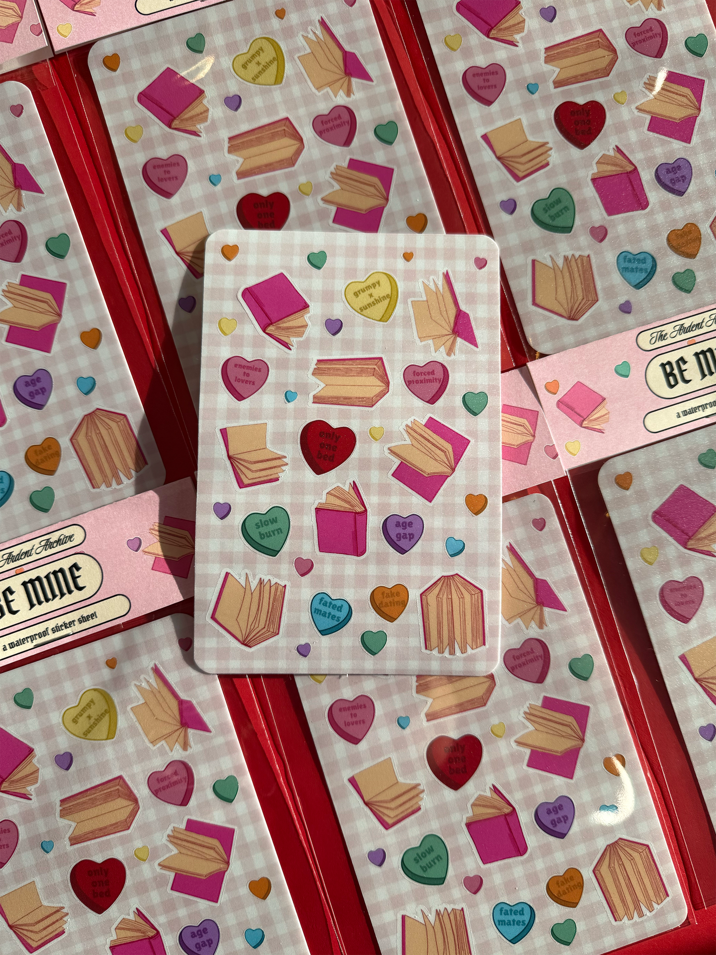 Six packaged sticker sheets with hearts and book designs on a checkered pink background arranged in a square, with an unpackaged sticker sheet askew on top.