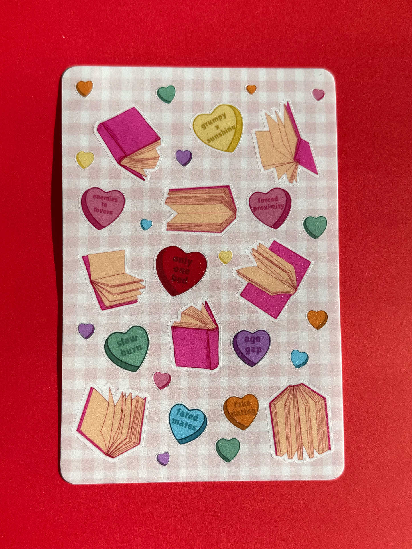 Unpackaged physical sticker sheet with hearts and book designs on a checkered pink background.