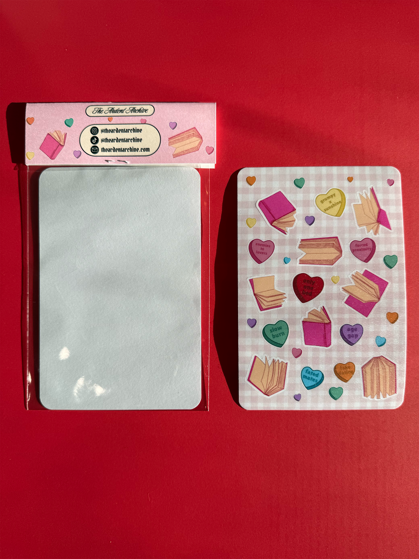 The back of one packaged sticker sheet, which is blank. And a second sticker sheet with hearts and book designs on a checkered pink background facing up.