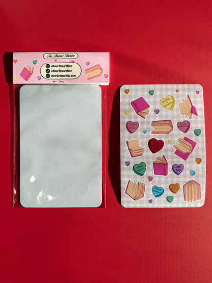 The back of one packaged sticker sheet, which is blank. And a second sticker sheet with hearts and book designs on a checkered pink background facing up.
