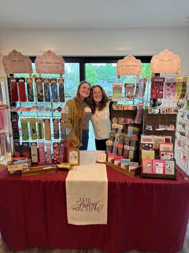 Two women standing behind a display table with various products and a sign reading 'The Ardent Archive'.