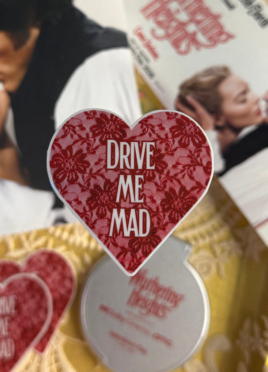 Heart-shaped sticker with 'Drive Me Mad' text on a blurred background of people and text.