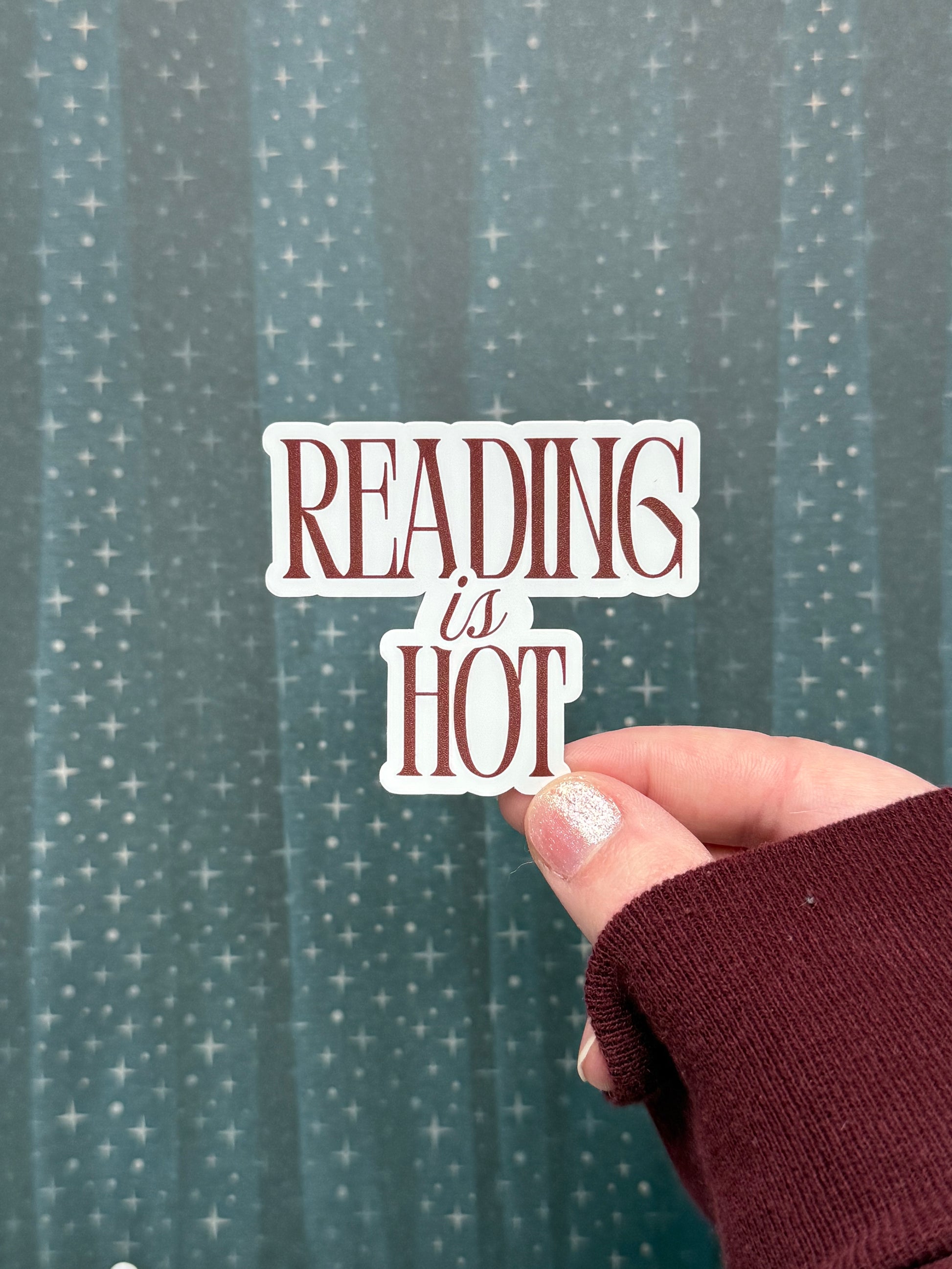 Hand holding a 'READING is HOT' sticker against a star-patterned curtain background