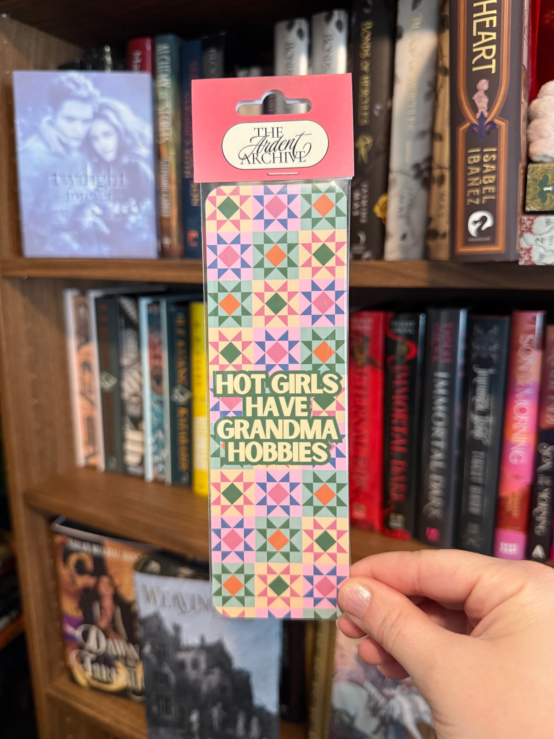 Hand holding a colorful bookmark with text in front of a bookshelf.