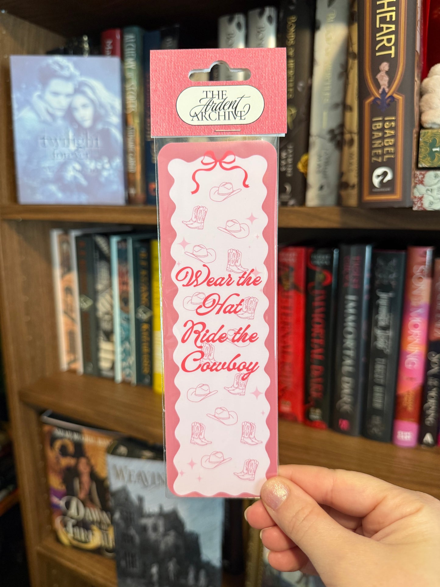 Pink bookmark with 'Wear the Hat, Ride the Cowboy' text held in front of a bookshelf.