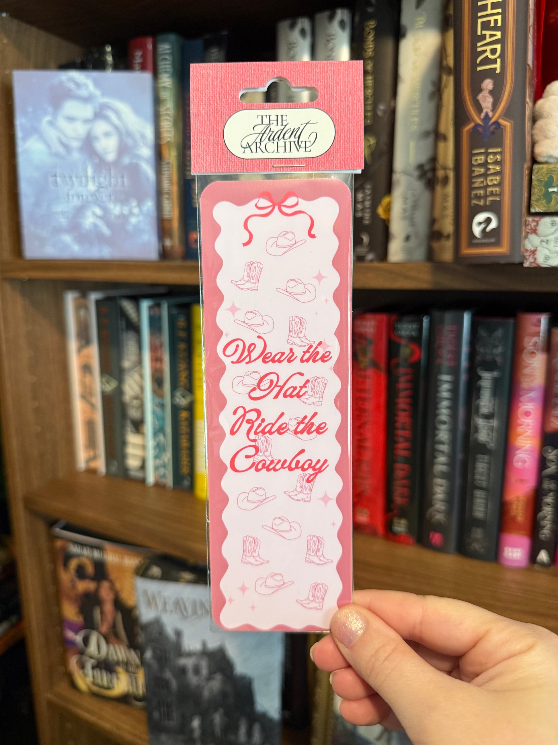 Pink bookmark with 'Wear the Hat, Ride the Cowboy' text held in front of a bookshelf.