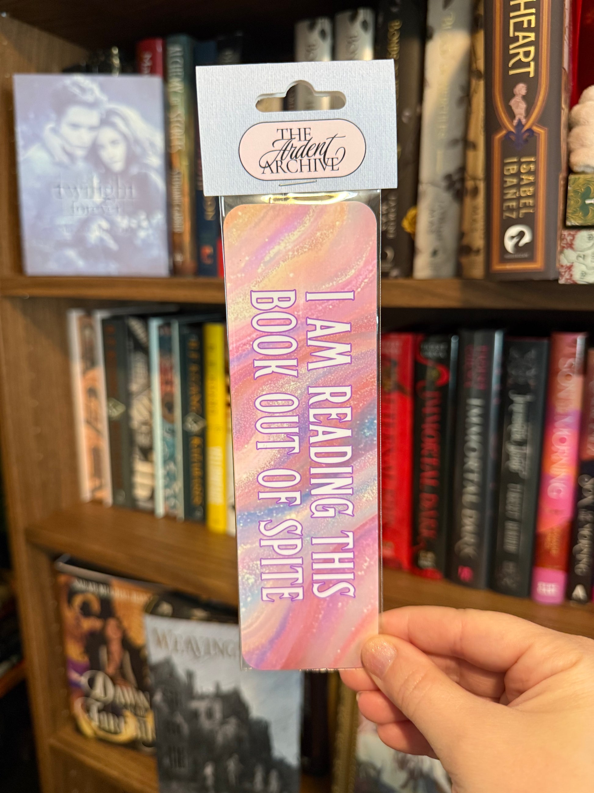 Bookmark with 'I am reading this book out of spite' text held in front of a bookshelf.