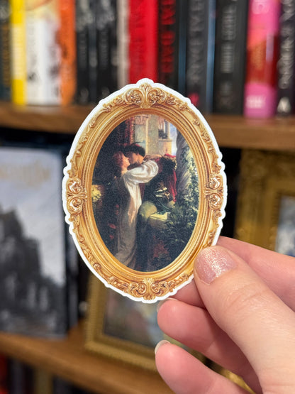 Decorative sticker with a gold frame and couple kissing held by a hand in front of a bookshelf.