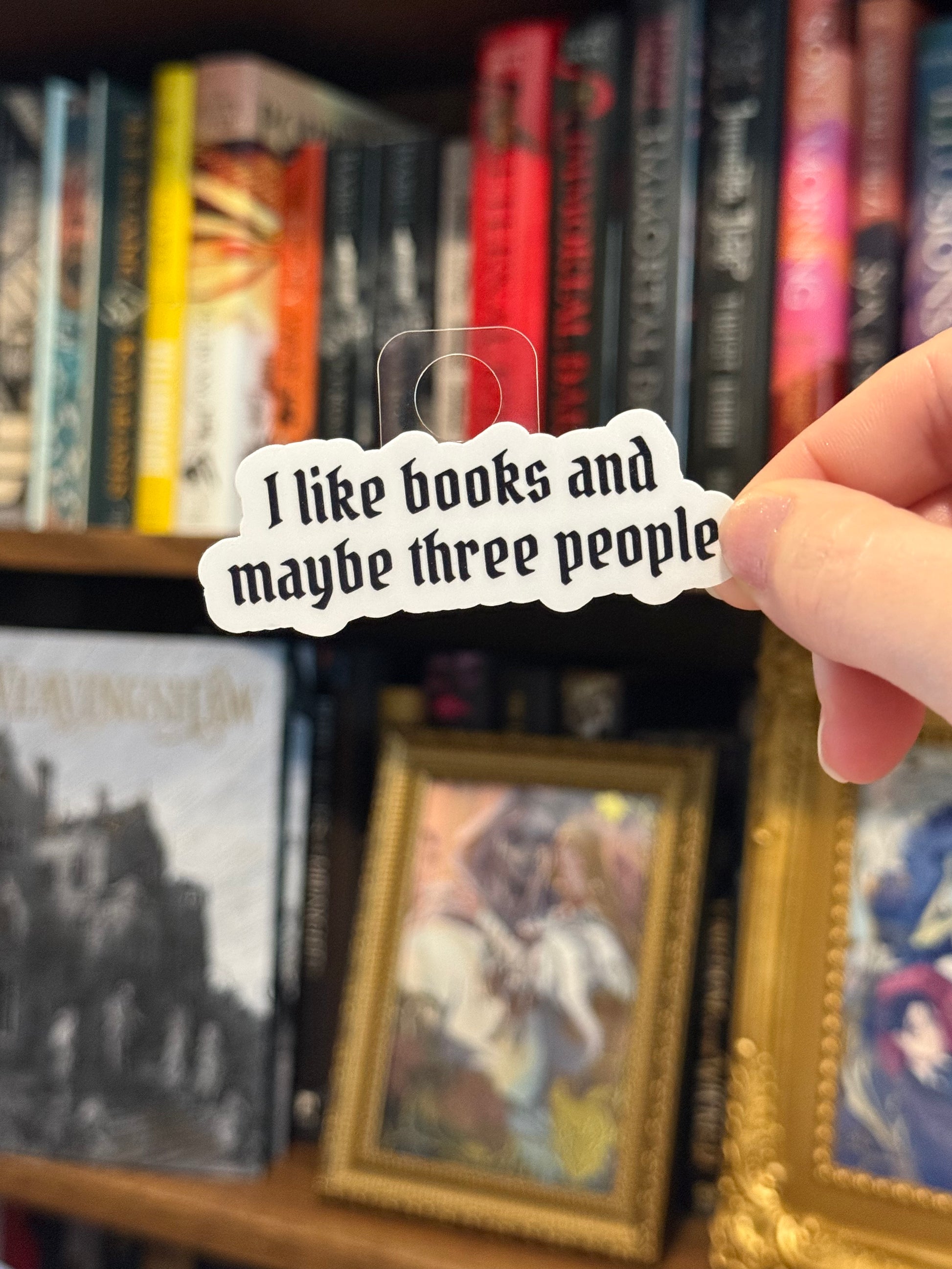 Person holding a sticker with text 'I like books and maybe three people' in front of a bookshelf.