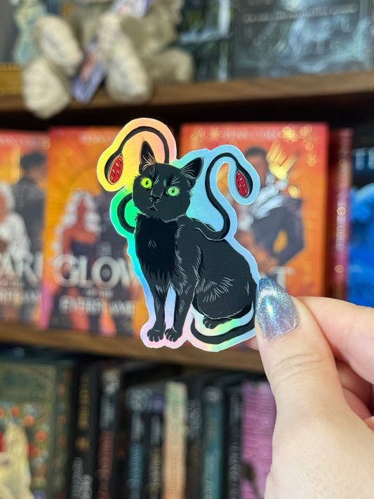 A hand with glittery nail polish holds a die-cut sticker of a black cat with colorful neon outlines, set against a blurred background of bookshelves.
