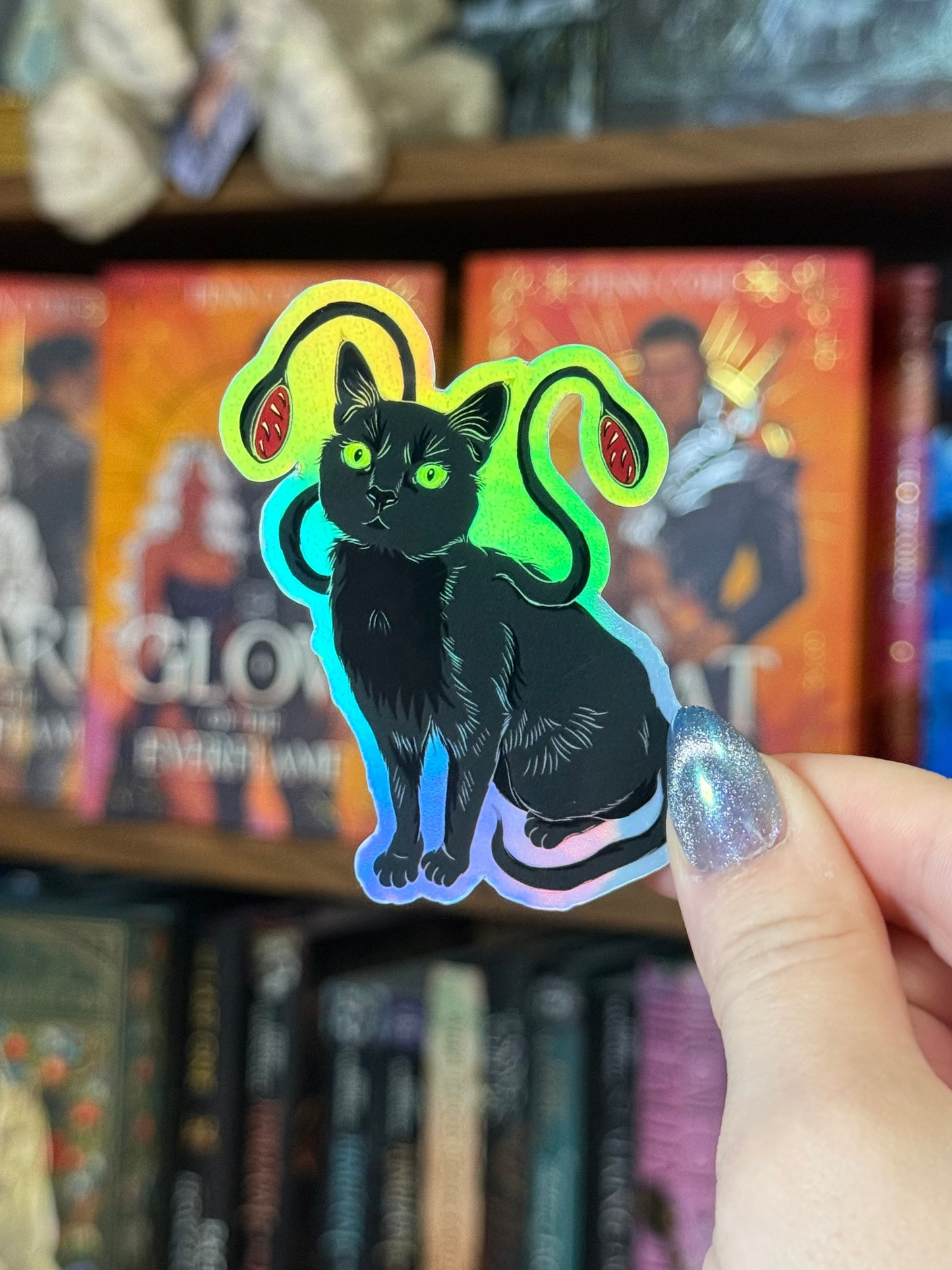 A hand with glittery fingernails holds a die-cut sticker of a black cat with glowing neon green and blue outlines in front of a blurred bookshelf.