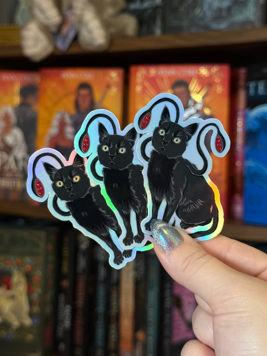 Three holographic stickers featuring black cats with red ears, held by a hand in front of a blurred bookshelf background.