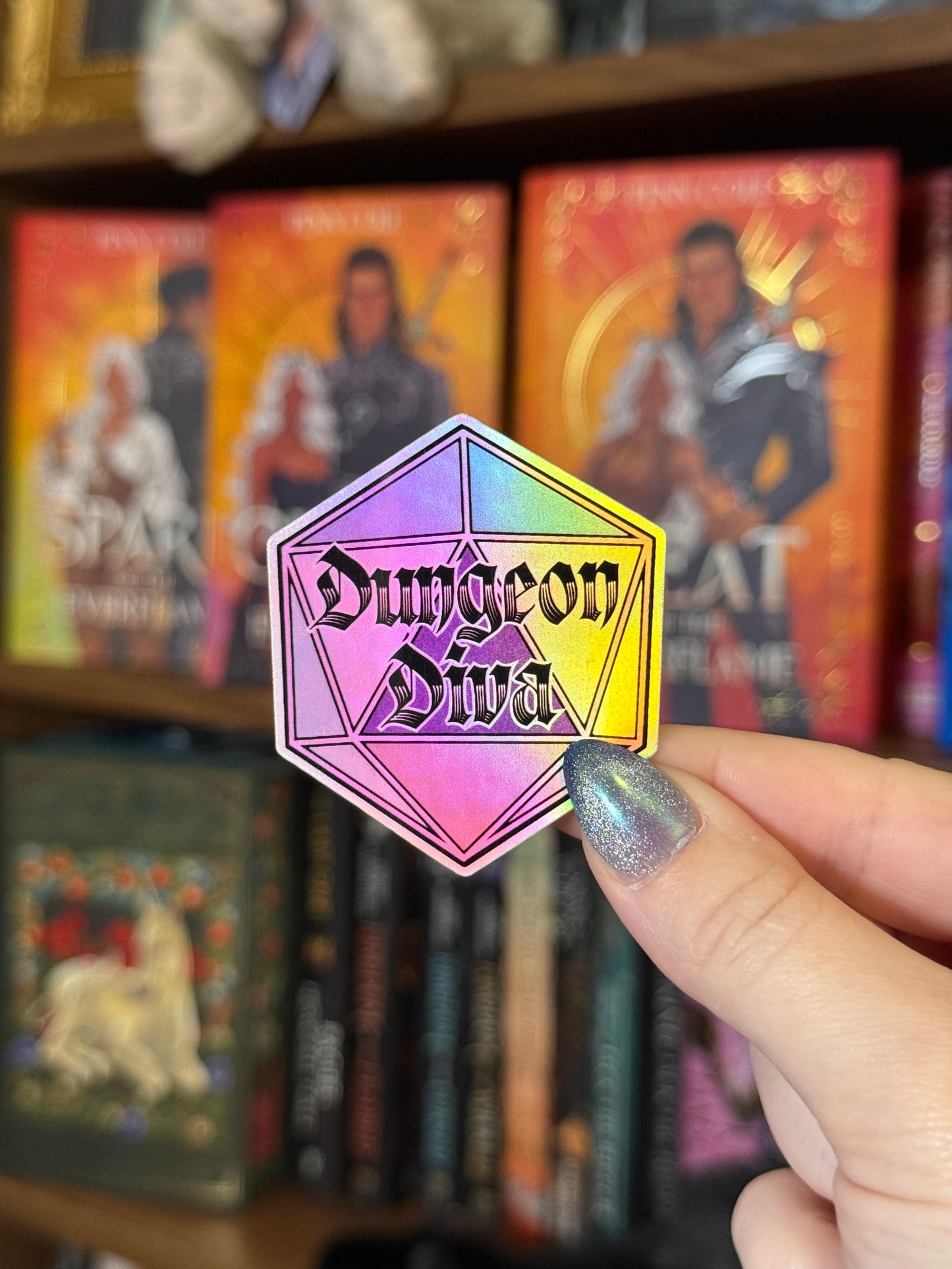 A hand with glittery silver nail polish holds a holographic hexagonal sticker featuring the text 'Dungeon Diva' in a black gothic font. The sticker has a rainbow iridescent finish shifting from pink to yellow.