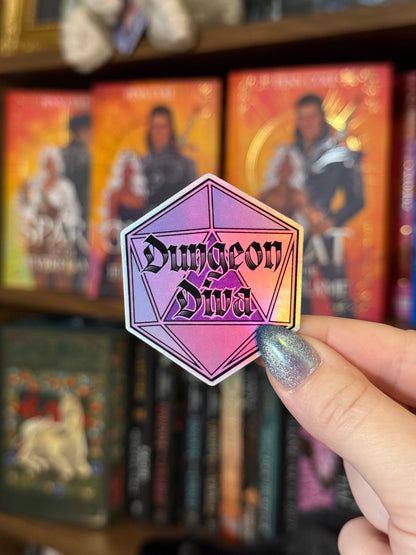 A hand with glittery silver nail polish holds a hexagonal sticker featuring a pink and purple 20-sided die (d20) design with the text 'Dungeon Diva' in black gothic lettering. The background is a blurred bookshelf filled with fantasy novels.