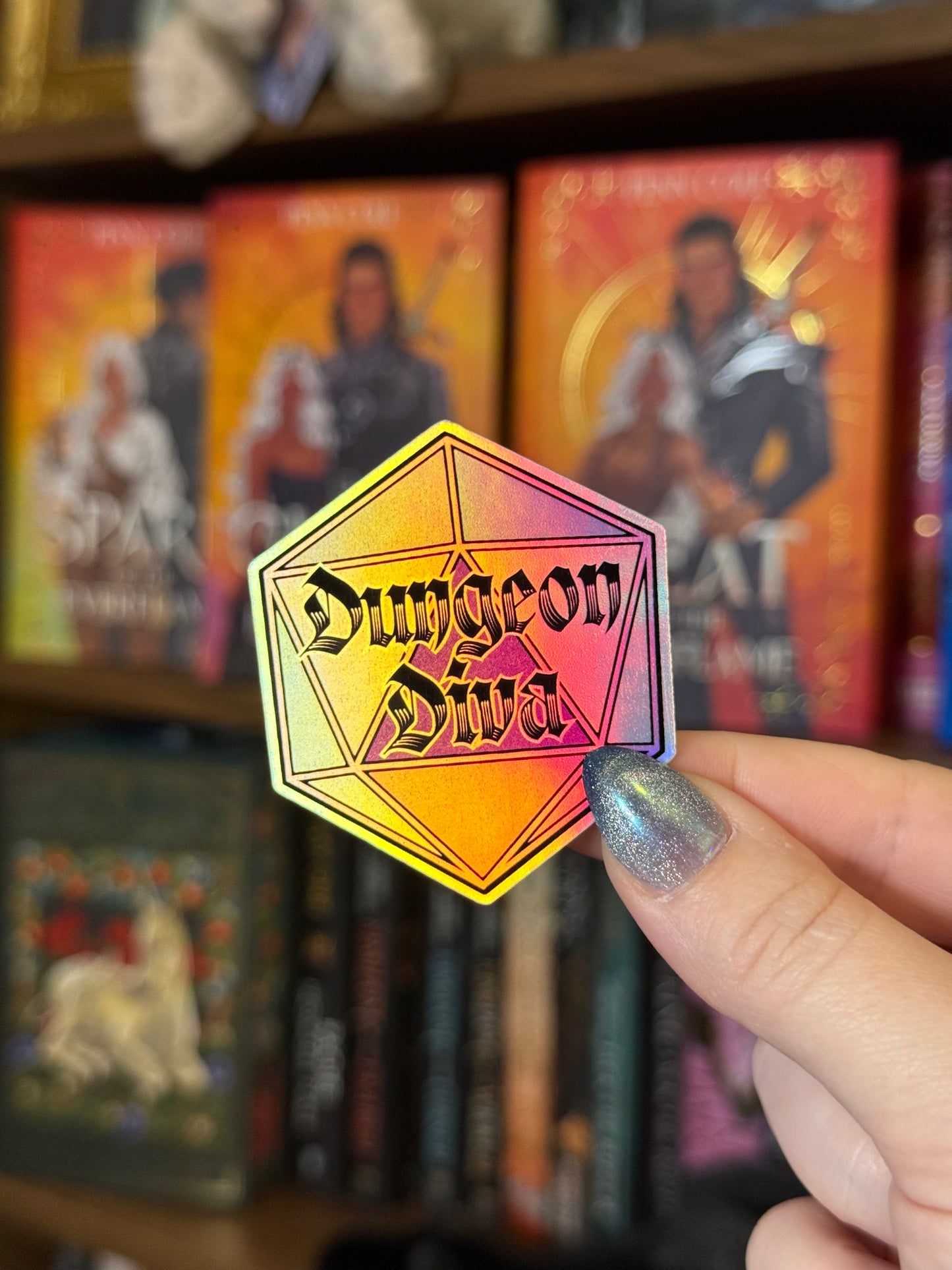 A hand with glittery silver nail polish holds a holographic hexagonal pin that reads 'Dungeon Diva' against a blurred background of fantasy bookshelves.