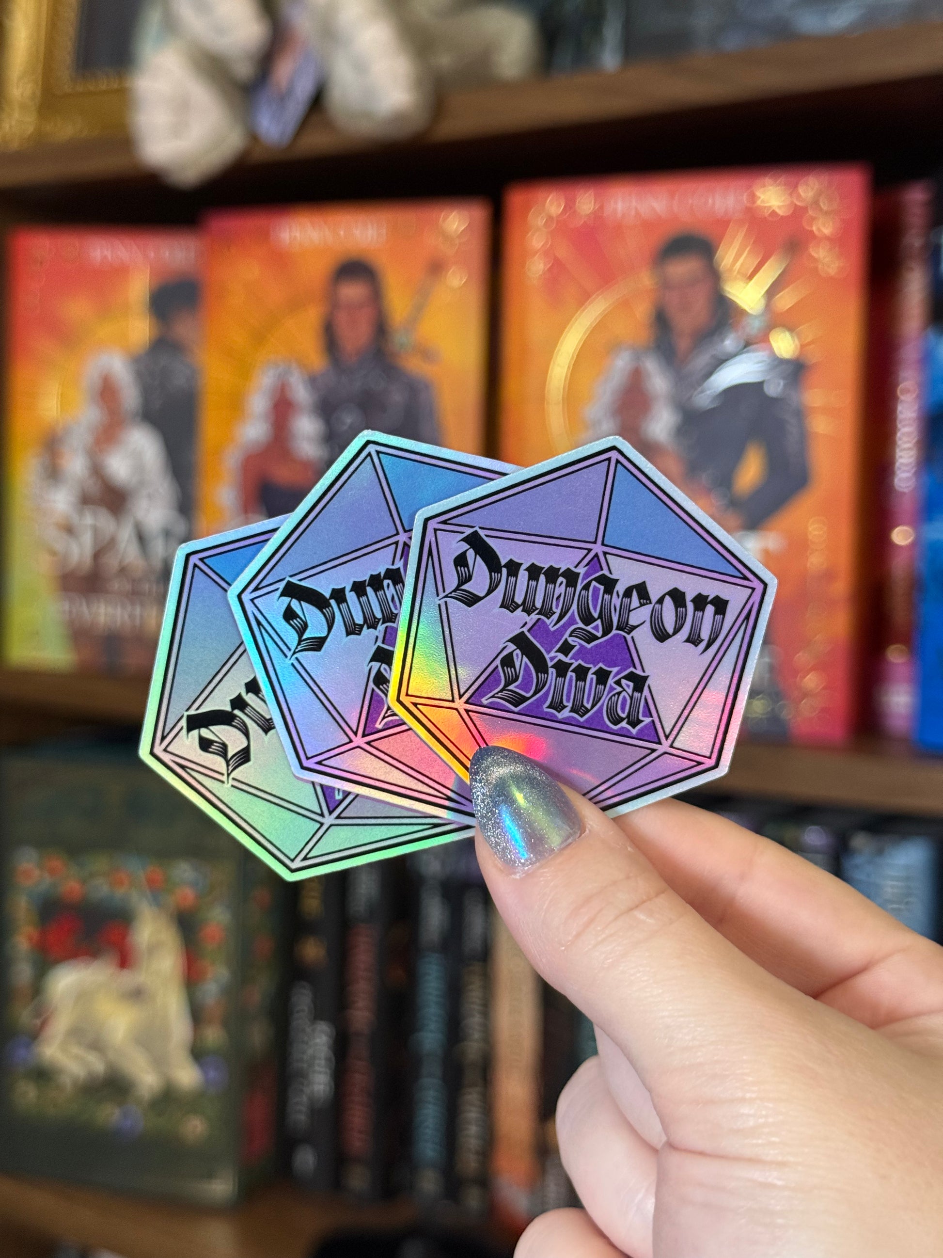 Three holographic diamond-shaped stickers reading 'Dungeon Diva' are held in a hand in front of a blurred bookshelf background.
