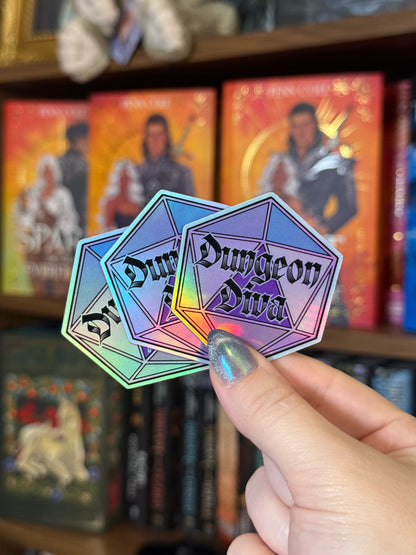 Three holographic diamond-shaped stickers reading 'Dungeon Diva' are held in a hand in front of a blurred bookshelf background.