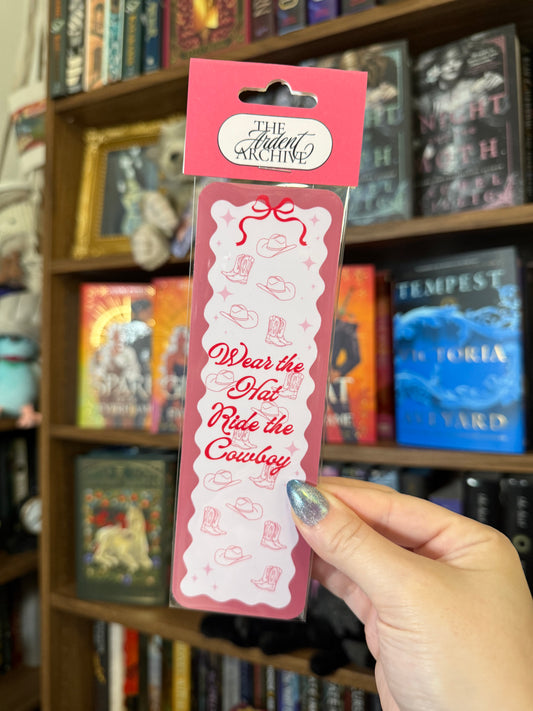 Pink bookmark being help up against bookshelf in background. Bookmark says "wear the hat, ride the cowboy"