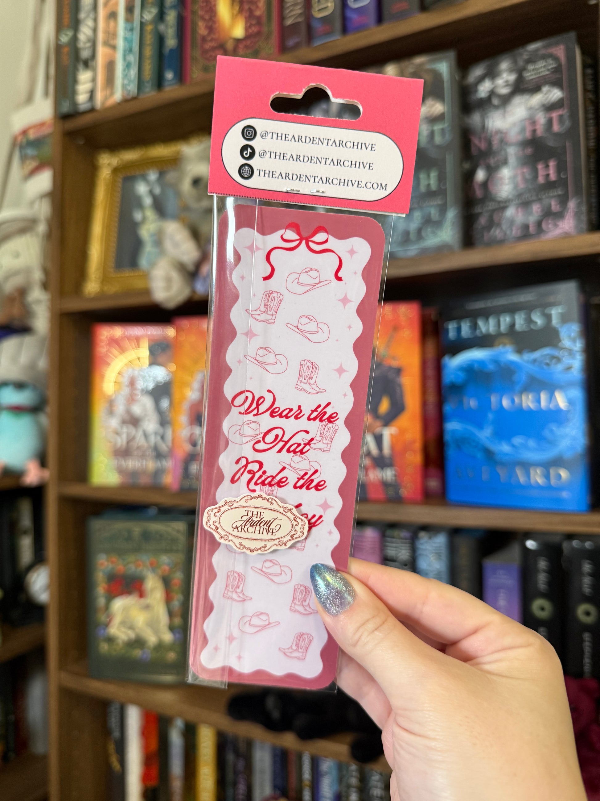 Pink bookmark being help up against bookshelf in background. Bookmark says "wear the hat, ride the cowboy"