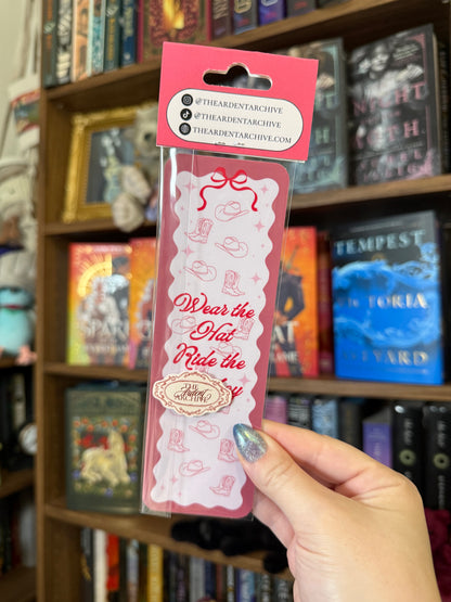 Pink bookmark being help up against bookshelf in background. Bookmark says "wear the hat, ride the cowboy"