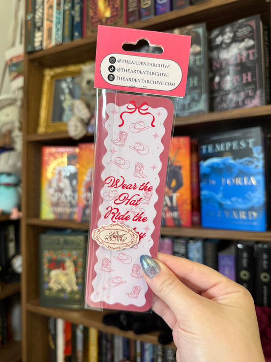 Pink bookmark being help up against bookshelf in background. Bookmark says "wear the hat, ride the cowboy"