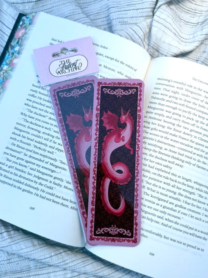 A decorative bookmark featuring a pink dragon on a dark background with ornate pink borders, placed on an open book with a pink hang tag that reads 'The Ardent Archive'.