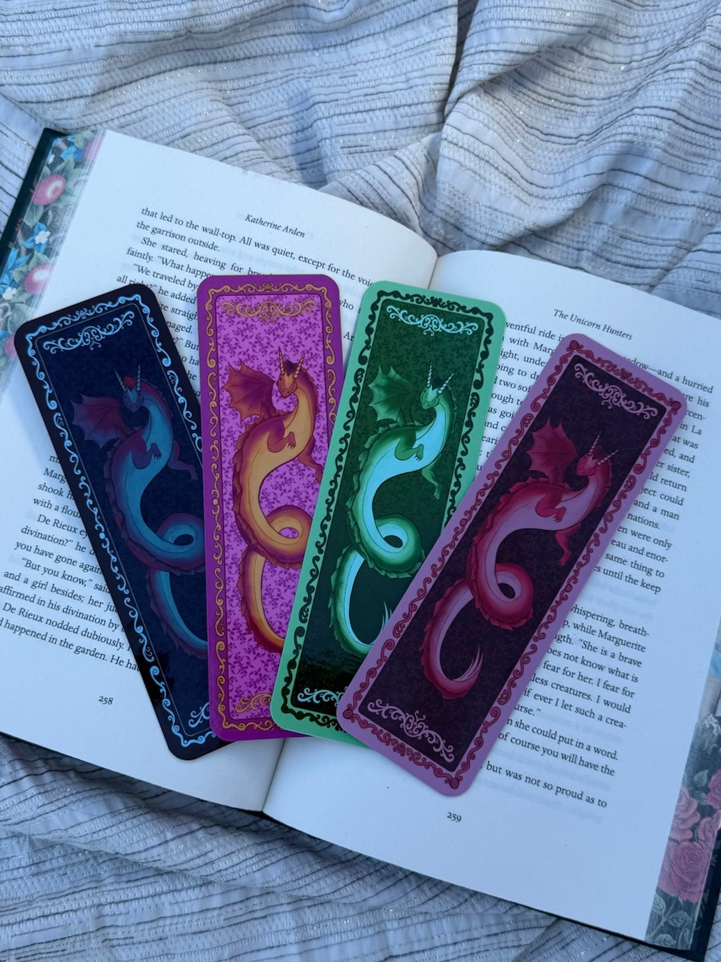 Four decorative dragon-shaped bookmarks in different colors (black, purple, green, and magenta) are arranged on an open book, set against a background of gray striped bedding.