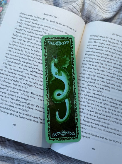 A green decorative book bookmark with a carved snake design and ornate borders, placed on an open book page.