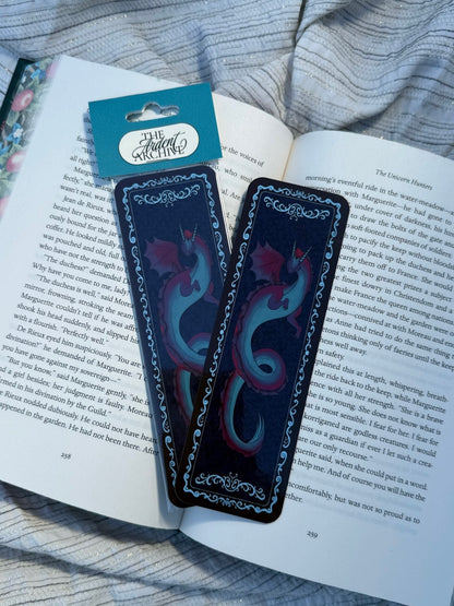 Two decorative book bookmarks shaped like dragons, resting on an open book. One bookmark features a red dragon with a teal body, and the other features a teal dragon with red wings.