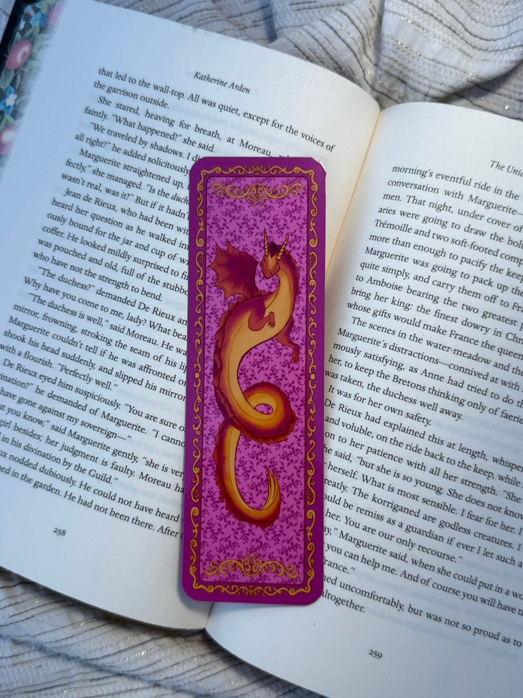A purple and gold dragon-themed bookmark resting on an open book page, with a cozy blanket in the background.