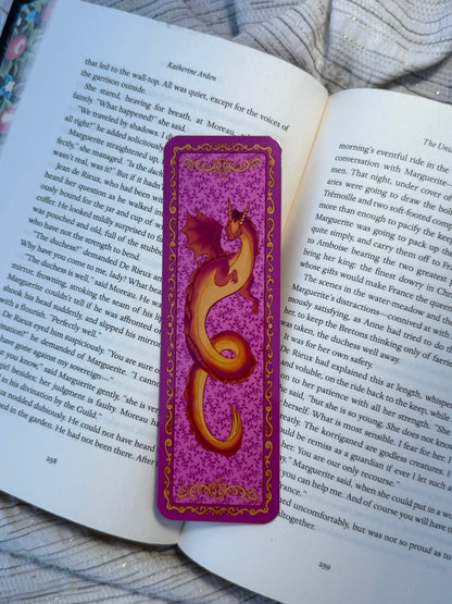 A purple and gold dragon-themed bookmark resting on an open book page, with a cozy blanket in the background.