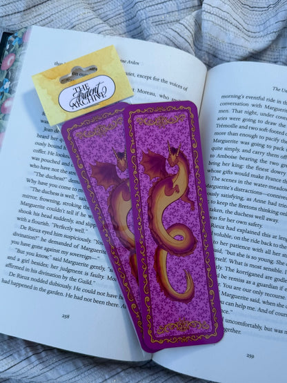 A purple and gold illustrated bookmark shaped like a stylized letter 'S' with a dragon motif, resting on an open book with a yellow 'The Great Reading' card attached.