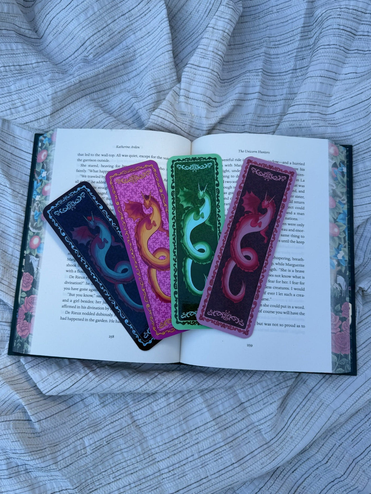 Three decorative book bookmarks with colorful dragon designs are laid across an open book on a gray striped fabric background.