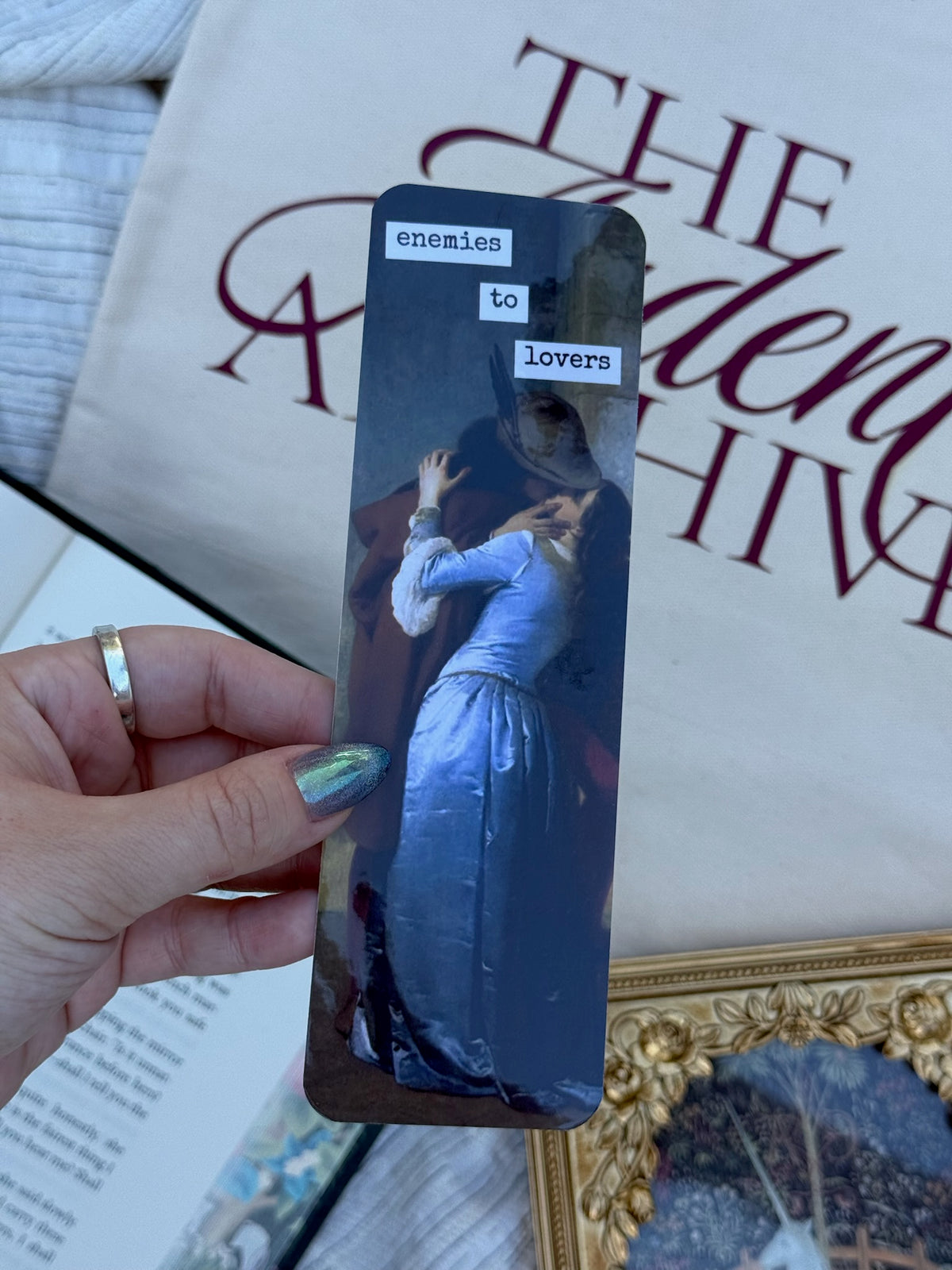 A hand with holographic nail polish holds a bookmark featuring a vintage painting of a woman in a blue dress kissing a man in a hat, with the words 'enemies to lovers' on top. A large tote bag with dark burgundy text reading 'The Ardent Archive' is in the background, along with books and a gold picture frame.
