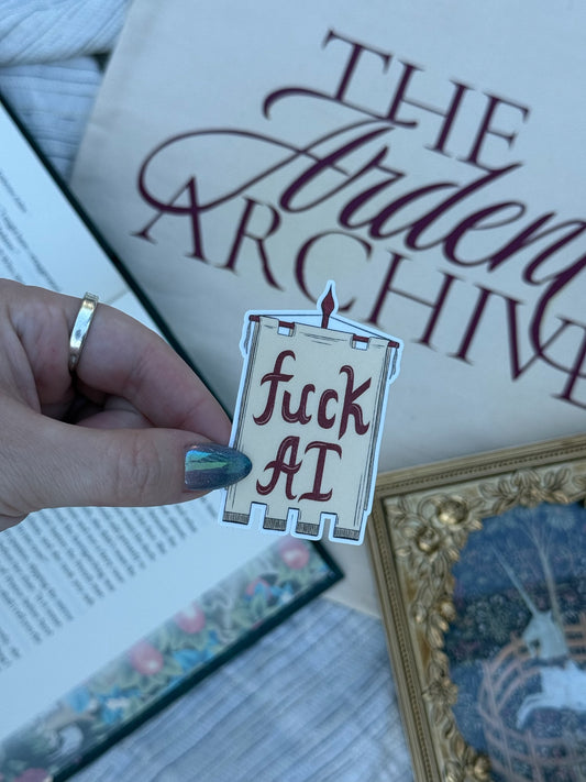 A hand with teal nail polish and a gold ring holds a die-cut sticker shaped like a parchment scroll that reads 'fuck AI' in dark red text. The background is a styled flat-lay featuring a book titled 'The Ardent Archive', a framed print, and a gray textured blanket.