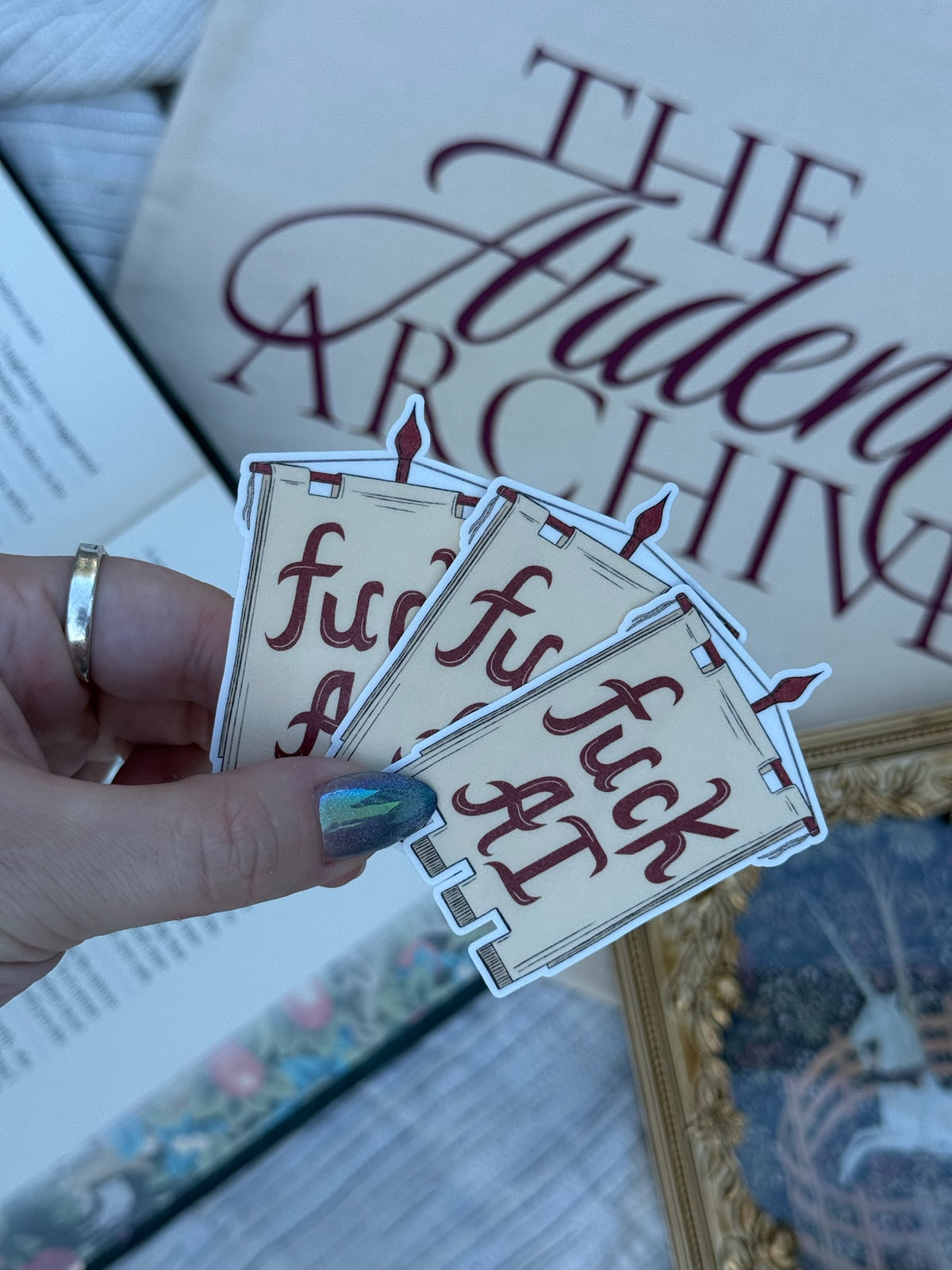 Three die-cut stickers featuring the phrase 'fuck AI' in dark red script on a beige banner design, held by a hand with iridescent nail polish. The background shows a bag with 'The Ardent Archive' on it and other vintage-themed items.