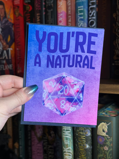 Hand holding a greeting card that says "youre a natural" with a D20 right underneath it in front of a bookshelf