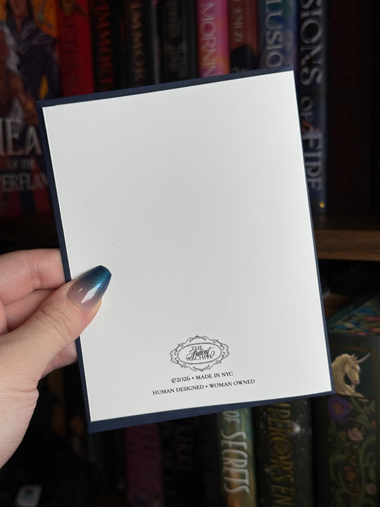 Hand holding a greeting card showing the back with a logo and information  front of a bookshelf
